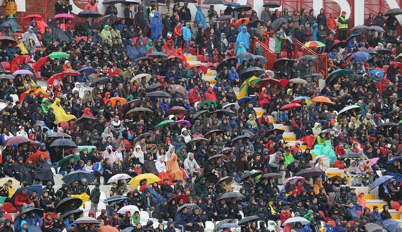 fans getting wet with umbrellas at football stadium without a roof