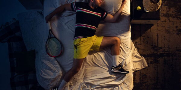 man asleep in bed with tennis kit on and tennis racquet